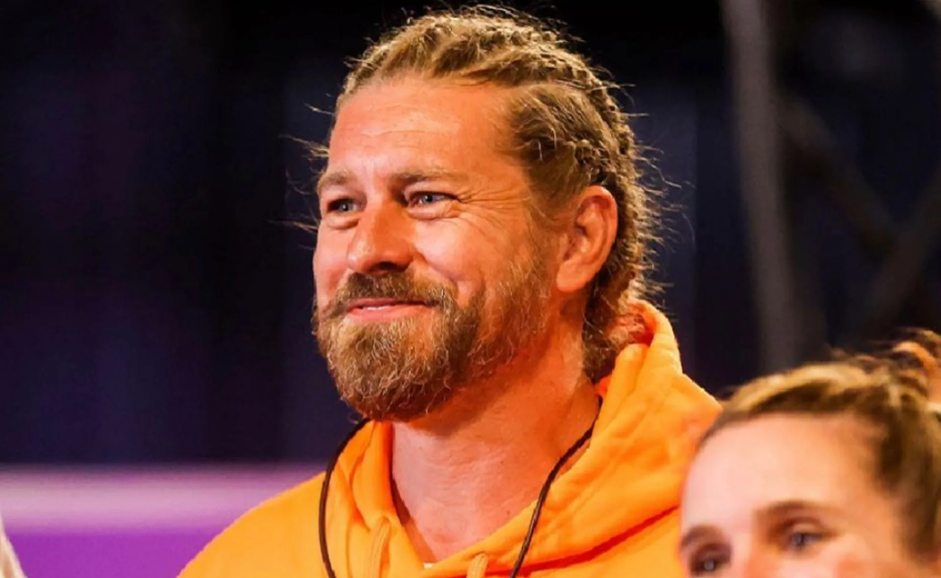 Man with braided hair and beard wearing an orange hoodie, smiling in a brightly lit setting.