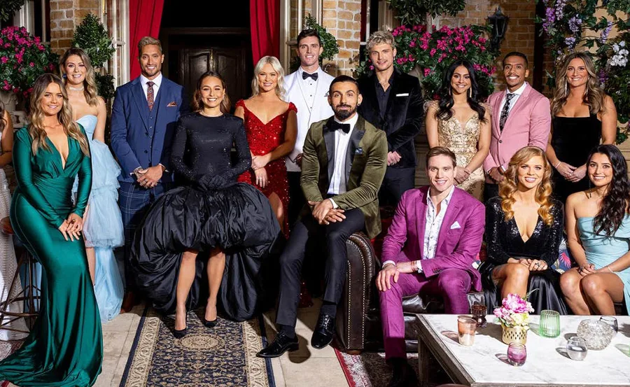 Contestants of the 2022 Australian Bachelorette pose in formal attire inside a room with floral decor.