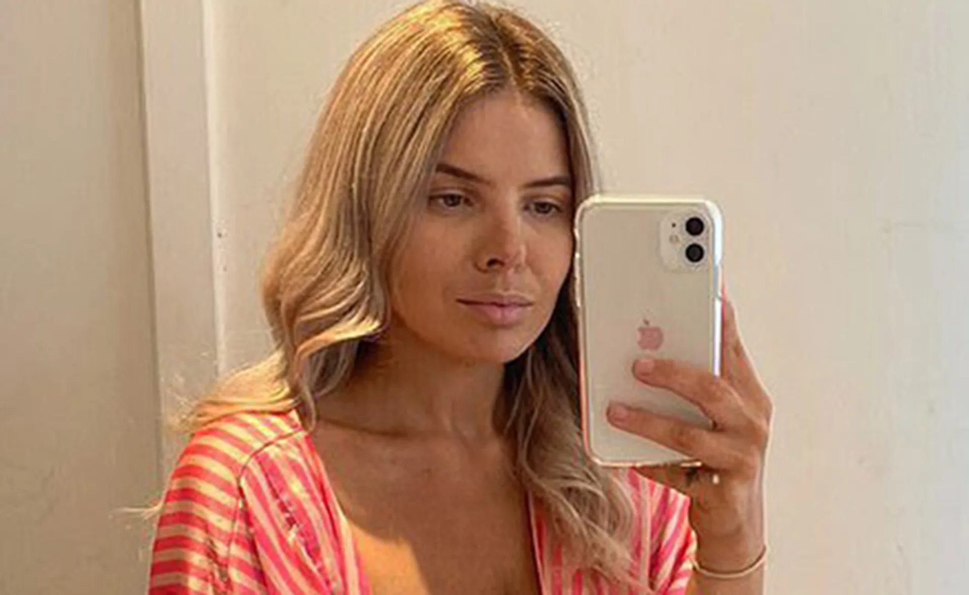 A person takes a selfie in a mirror, wearing a pink-striped top, holding a white smartphone.