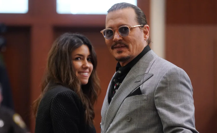Two people smiling, one in a gray suit with glasses, the other with long brown hair, inside a room with wooden walls.