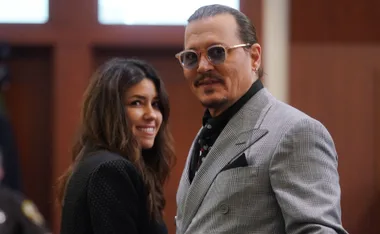 Two people smiling, one in a gray suit with glasses, the other with long brown hair, inside a room with wooden walls.