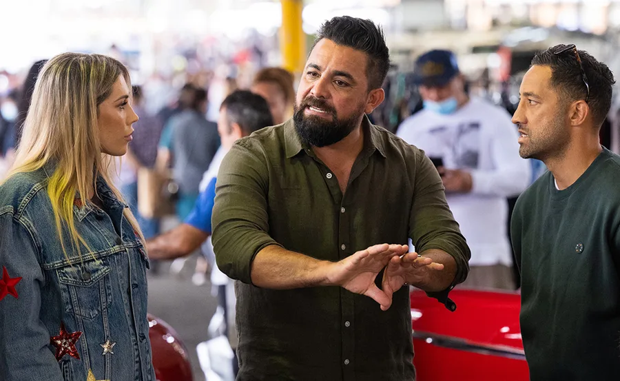 Three people engaged in a conversation at a busy indoor market setting.