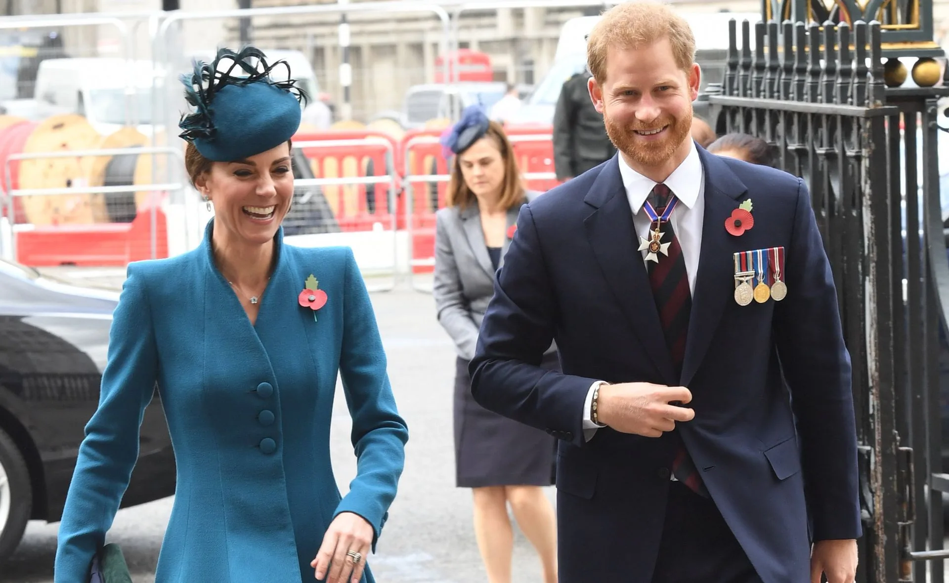 Two people smiling outdoors, dressed formally, wearing poppy pins and medals, with another person in the background.