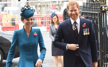 Two people smiling outdoors, dressed formally, wearing poppy pins and medals, with another person in the background.