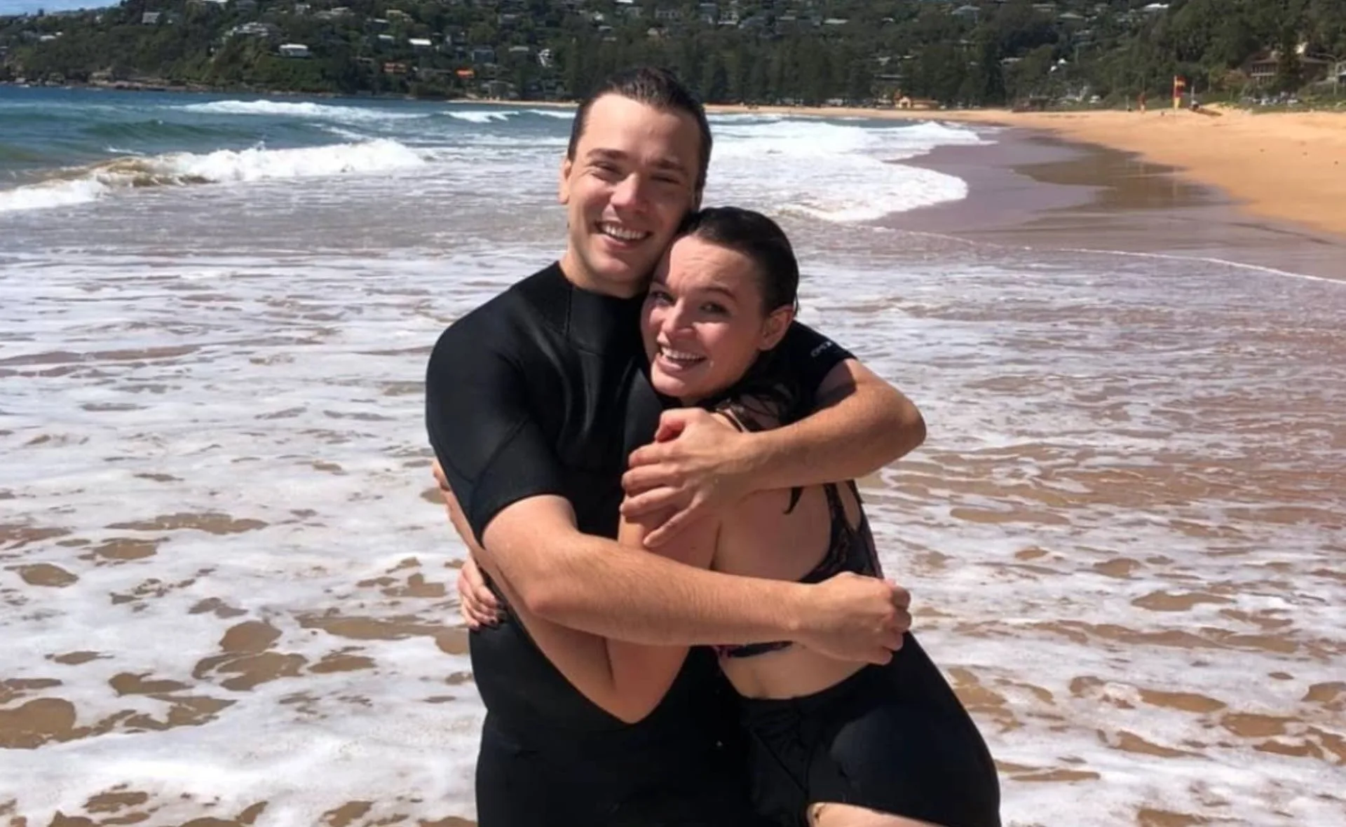 Two people smiling and hugging on a sunny beach, with waves in the background.