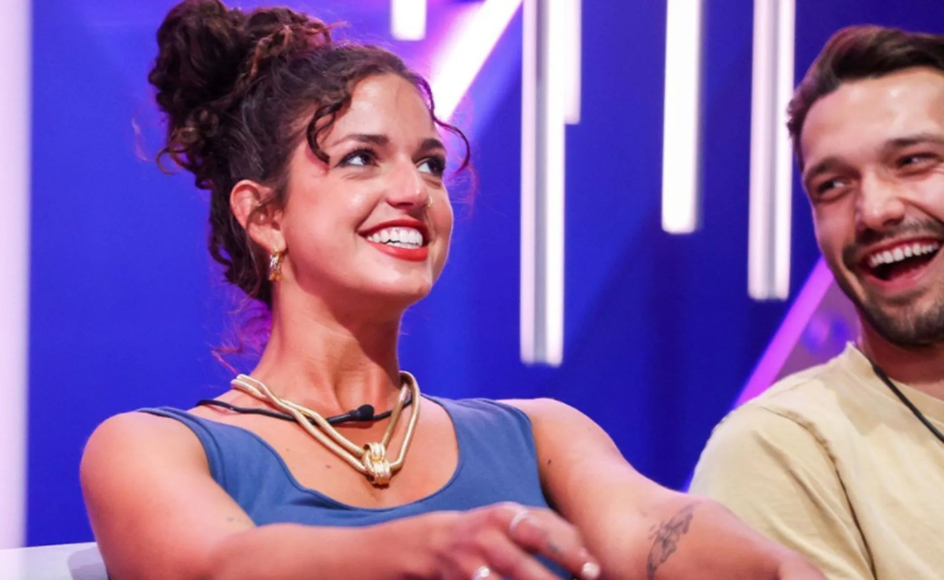 Two people smiling and sitting closely on a brightly lit TV set, engaged in conversation.