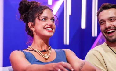 Two people smiling and sitting closely on a brightly lit TV set, engaged in conversation.