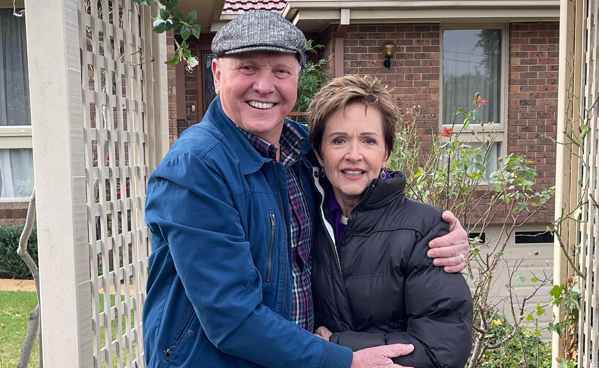 A smiling couple embraces in front of a brick house with a garden.