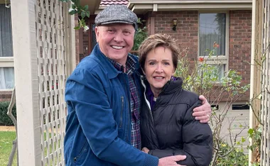 A smiling couple embraces in front of a brick house with a garden.