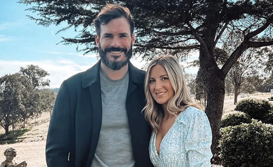 A couple smiling and posing outdoors, surrounded by trees and greenery.