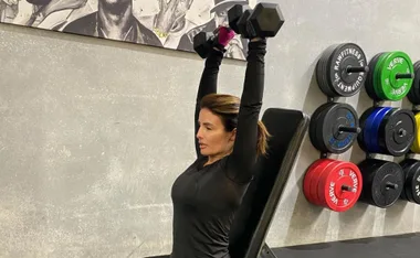 A woman in a gym performing a shoulder press with dumbbells, seated on a bench next to weight plates.