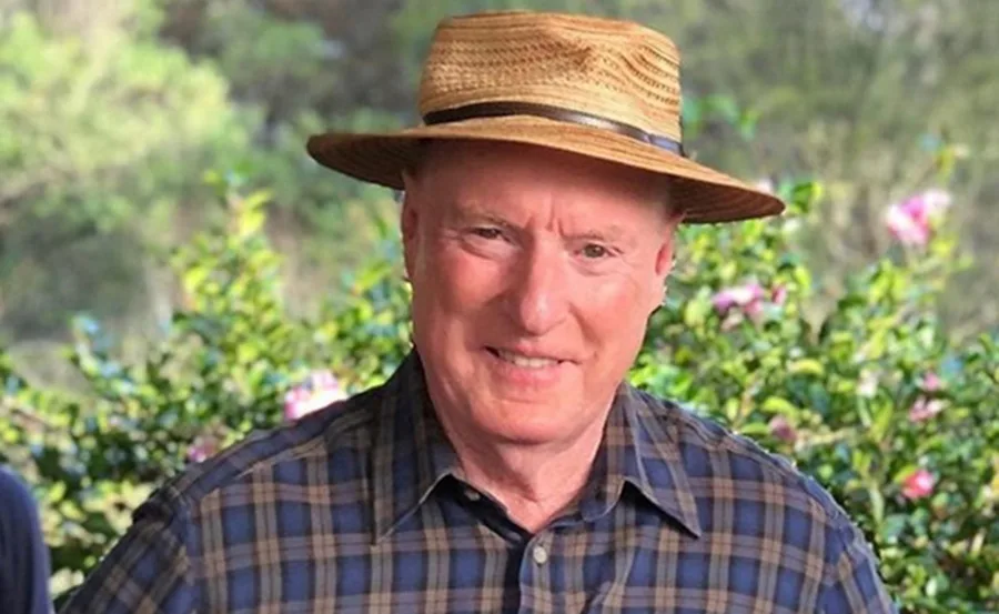 Man in a plaid shirt and straw hat smiling, outdoors with greenery in the background.