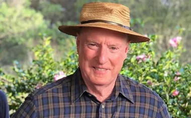 Man in a plaid shirt and straw hat smiling, outdoors with greenery in the background.