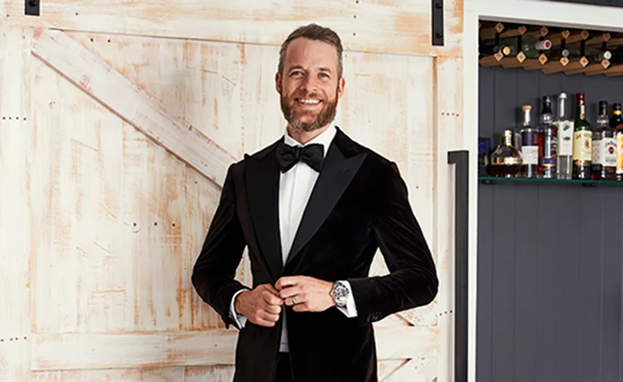 Man in a black tuxedo with a bow tie, smiling and buttoning jacket in front of a wooden door with bottles on a shelf behind.
