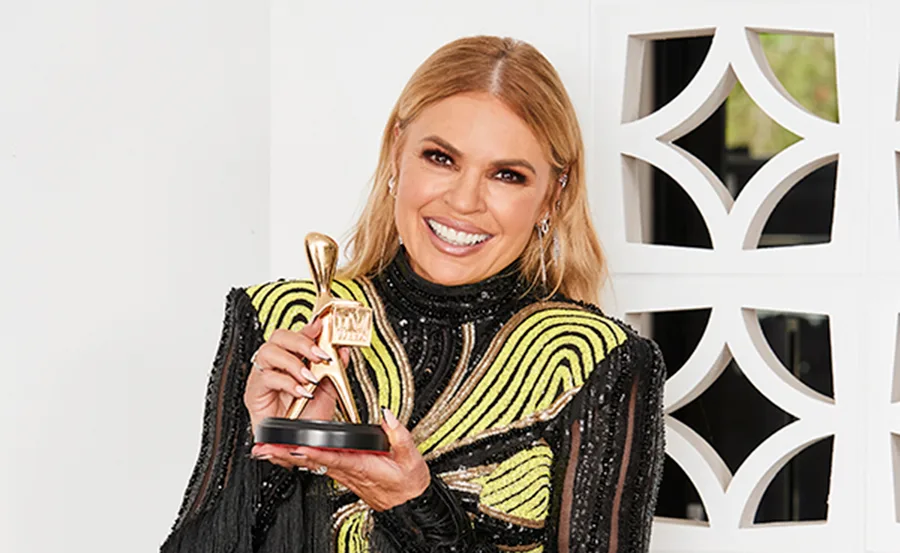 A woman smiles while holding a gold Logie award, wearing a black and yellow patterned dress.