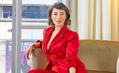 Person in a red suit sitting on a sofa, posing confidently near a window.