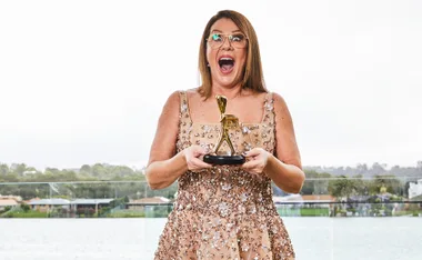 A woman in a sequined dress, holding a Logie award, stands with an excited expression against a river backdrop.