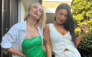 Two women posing outdoors, one in a green dress with a white shirt, the other in a white outfit with red lipstick.