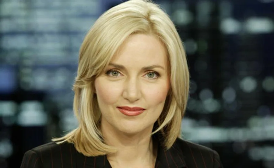 Blonde woman with short hair and a black jacket, smiling slightly, against a blurred background.