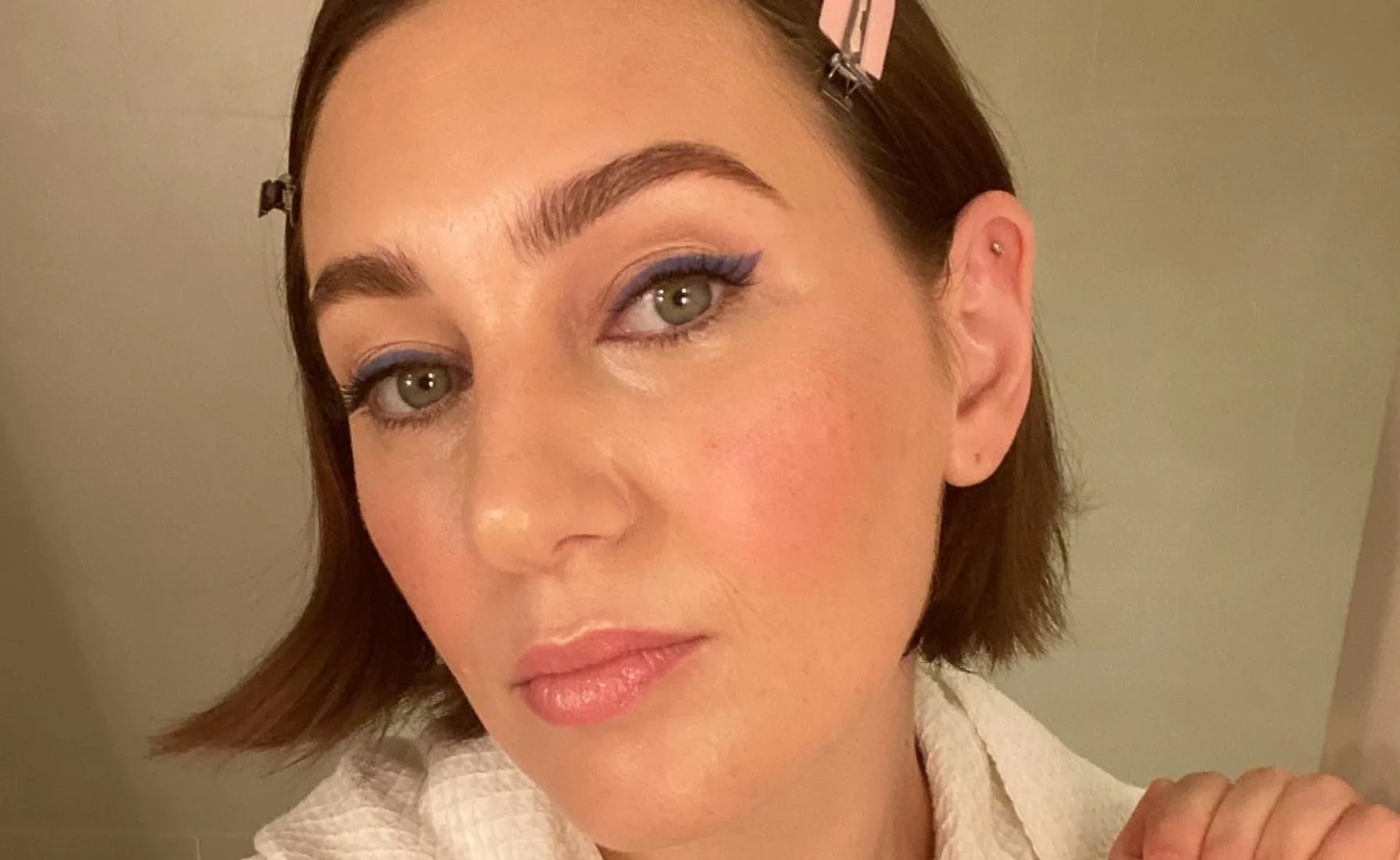 Woman with short hair, makeup, and pink hair clips, poses for a selfie in a bathroom setting.