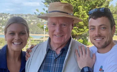 Three people smiling outdoors, with a scenic landscape background. The person in the center wears a hat.
