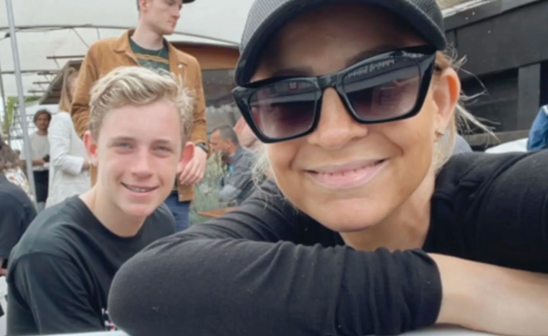 Woman in sunglasses and cap takes a selfie with a smiling boy at an outdoor gathering.