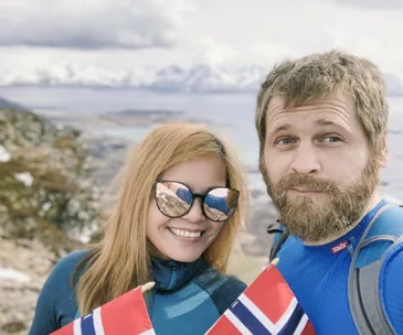 Happy couple with Norwegian flags hiking in snowy mountains, bright and cloudy sky in background.