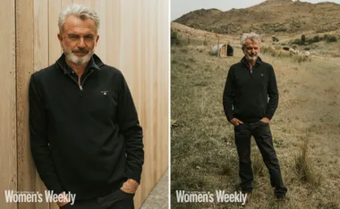 Man with gray hair and beard, wearing a black sweater, leans against a wooden wall and stands in a field with hills behind.