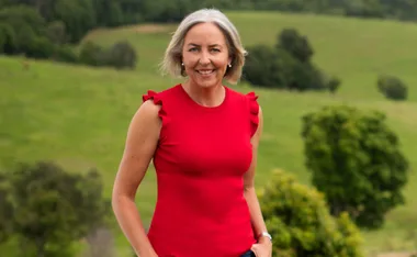 A woman in a red shirt smiles outdoors with a green landscape background.