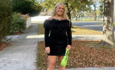 A woman in a black off-shoulder dress holding a green purse on a leafy sidewalk.