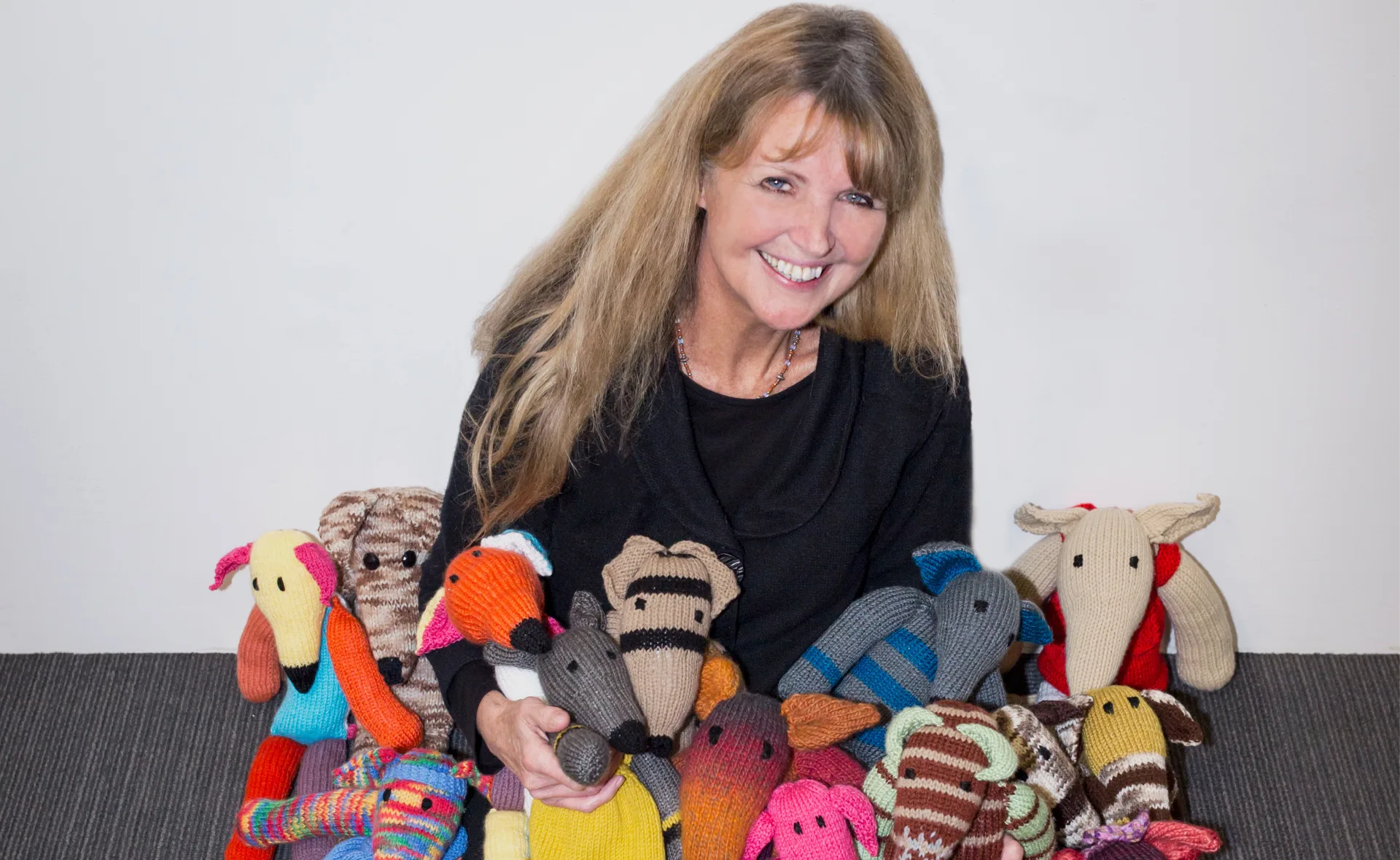 A woman smiles, surrounded by colorful knitted greyhound toys.