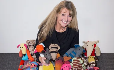 A woman smiles, surrounded by colorful knitted greyhound toys.