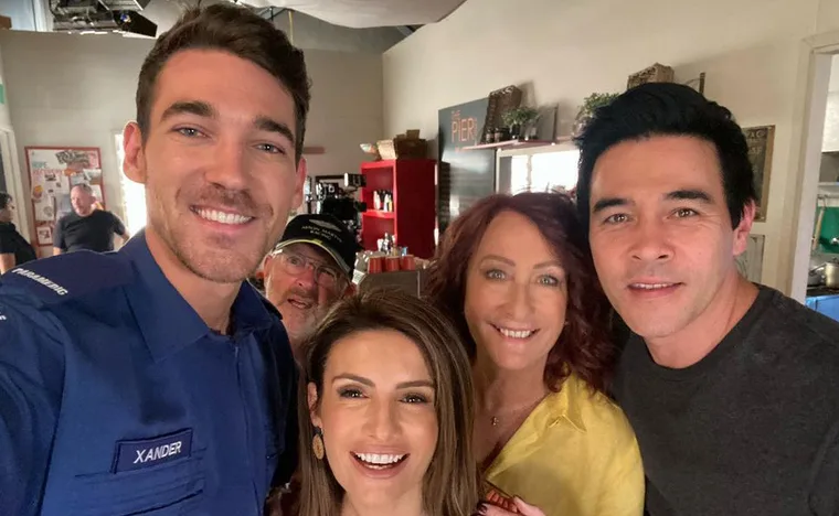 "Behind the scenes on Home and Away set, five cast members smiling together, one in a paramedic uniform labeled 'Xander'."