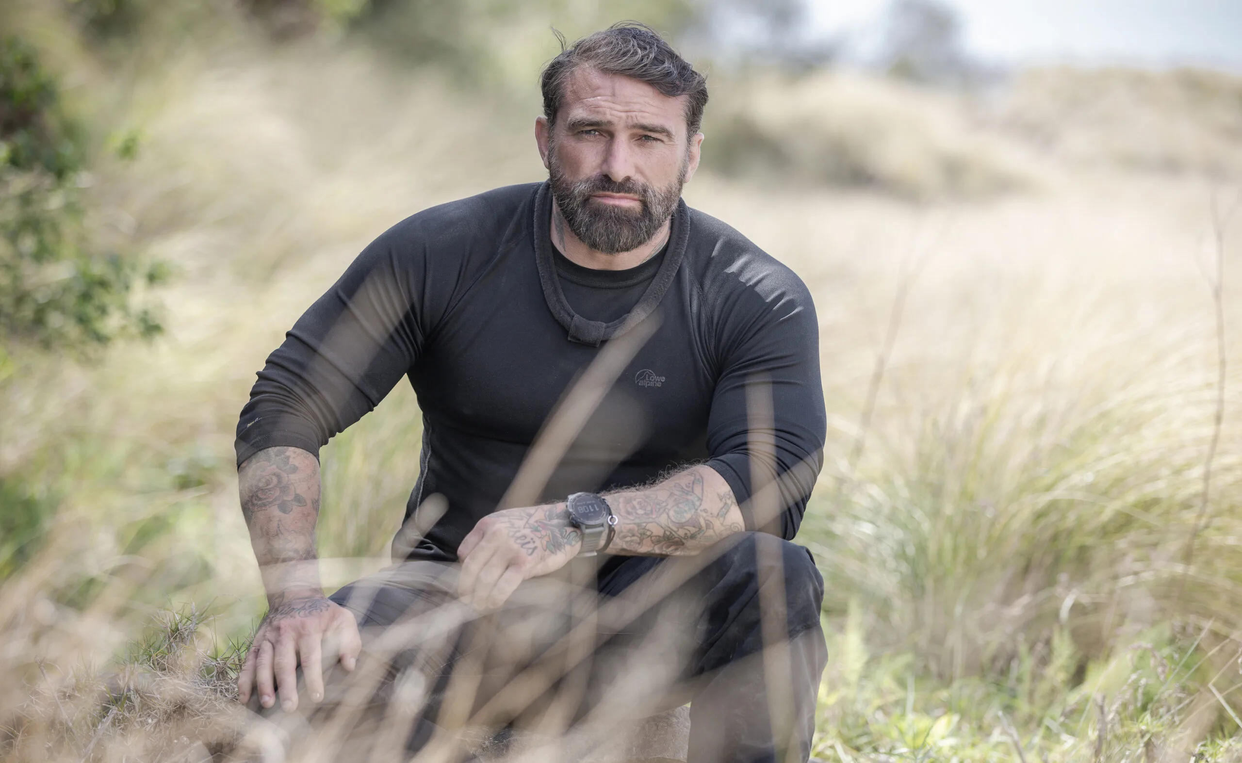 Man with tattoos in a black shirt sitting in a field of tall grass.