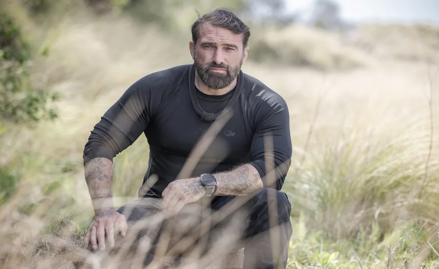 Man with tattoos in a black shirt sitting in a field of tall grass.