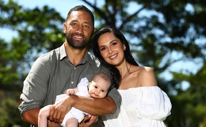 Family portrait of a couple holding their baby, standing outside with trees in the background.