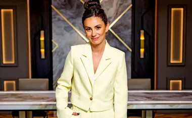 A woman in a light yellow suit stands confidently in a stylish interior with geometric decor.