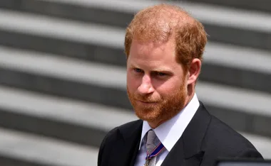 Prince Harry in a suit with a medal, standing outdoors against a staircase background.
