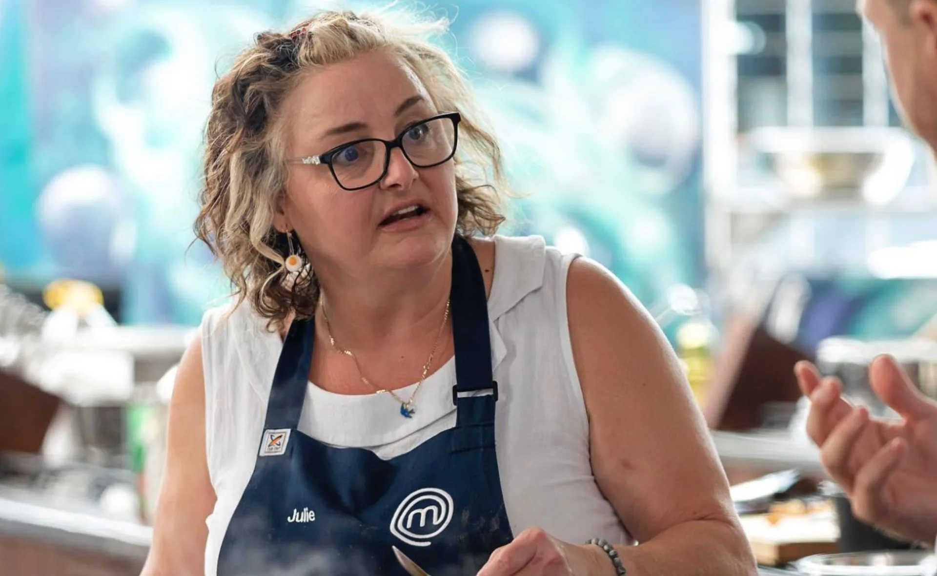 Woman with curly hair and glasses, wearing a MasterChef apron, looking thoughtful in a kitchen setting.