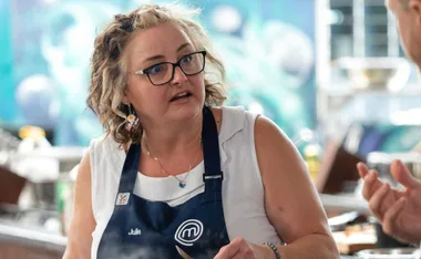 Woman with curly hair and glasses, wearing a MasterChef apron, looking thoughtful in a kitchen setting.
