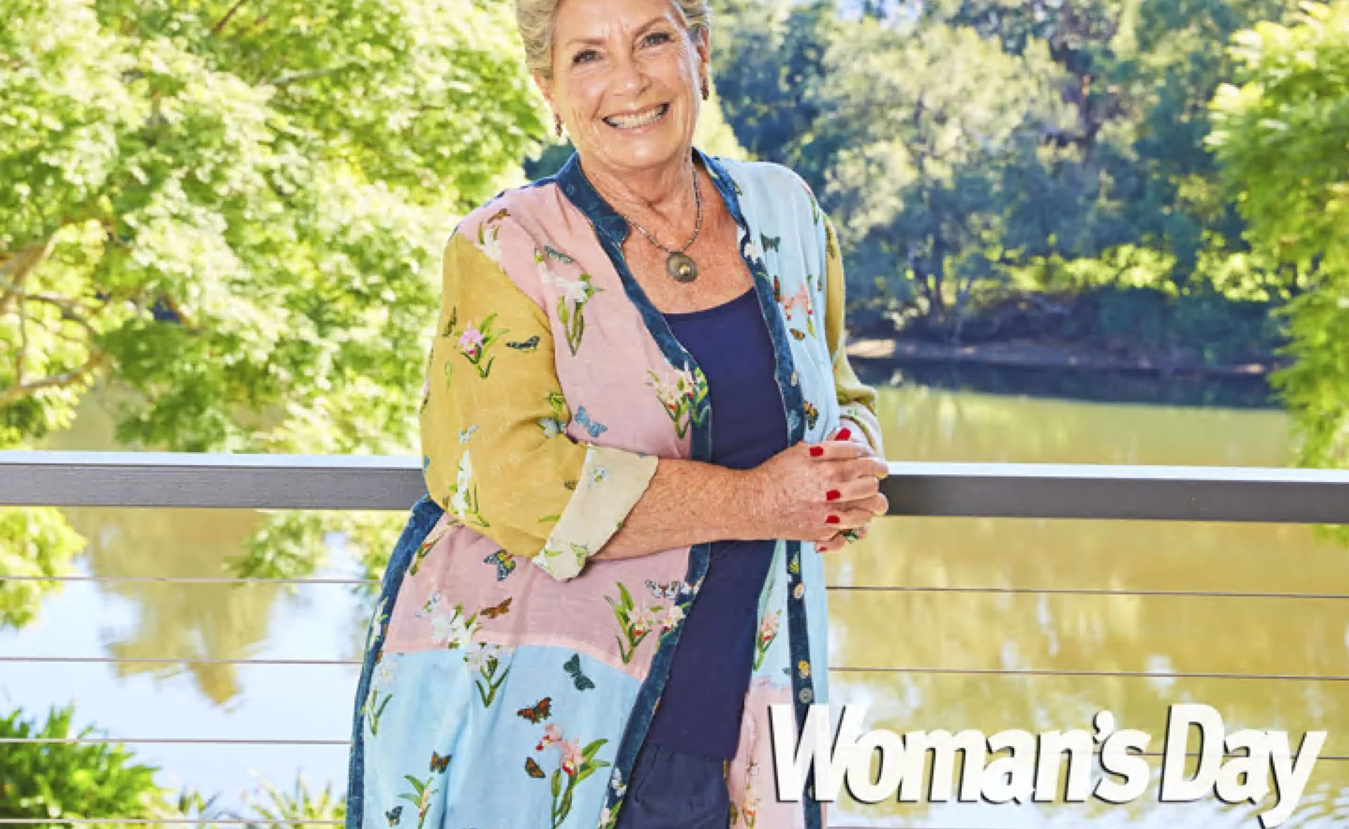 A woman smiling on a balcony, wearing a floral patterned jacket, with a scenic view of trees and a river.