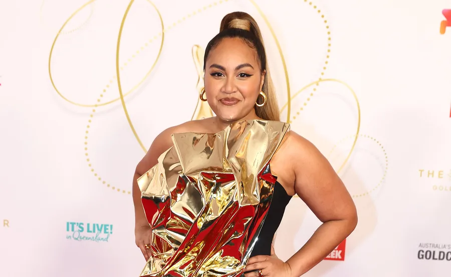 Jessica Mauboy on the red carpet in a striking gold and black dress, smiling confidently.