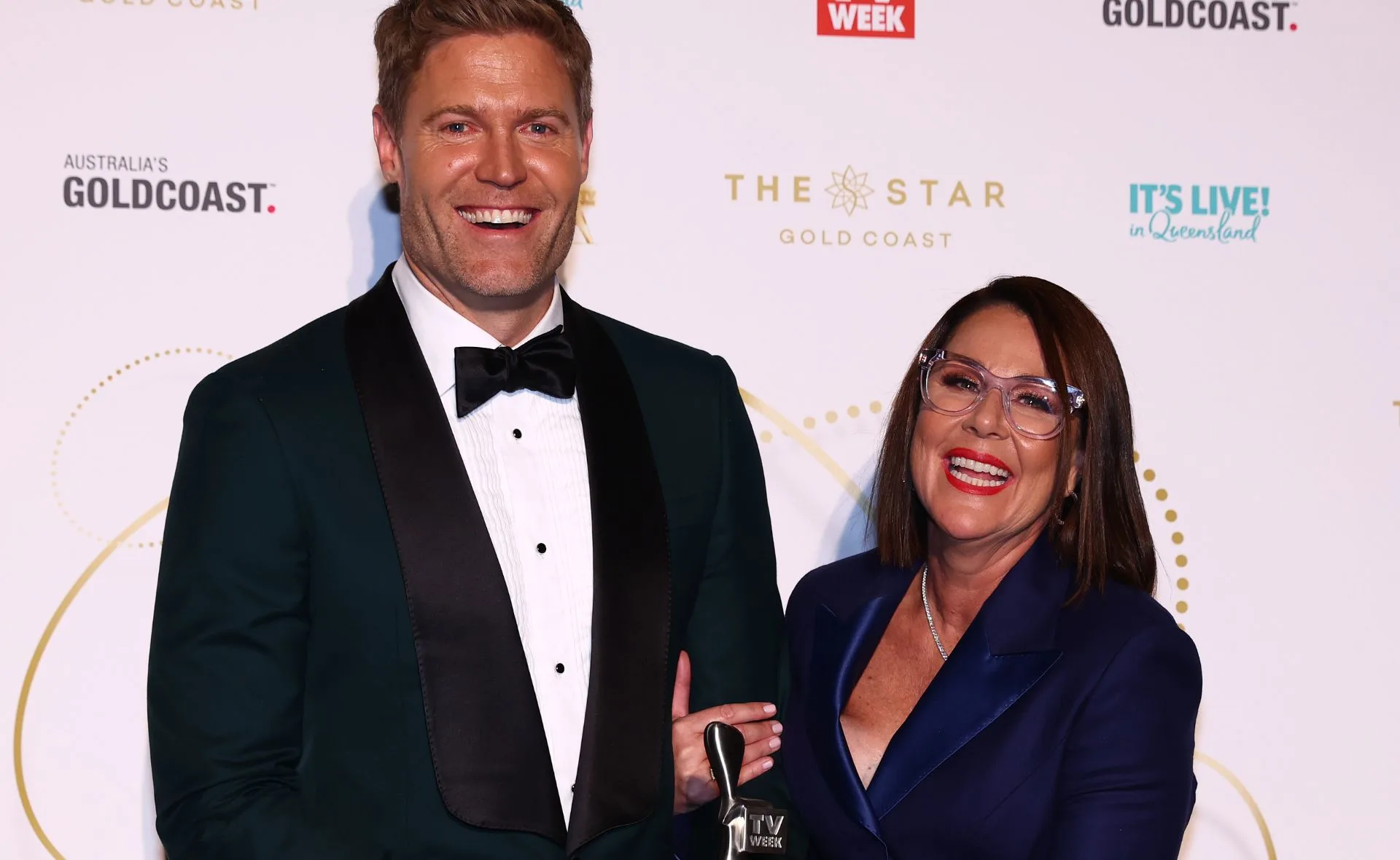 Two people smiling, dressed in formal attire at a red carpet event, with one holding an award statue.