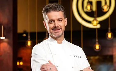 Chef in a white coat standing in front of a blurred background with hanging lights and a circular logo.