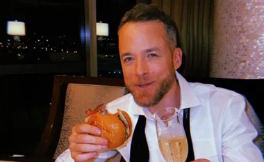 Man in a white shirt holds a burger and a glass of champagne, smiling at an after-party.