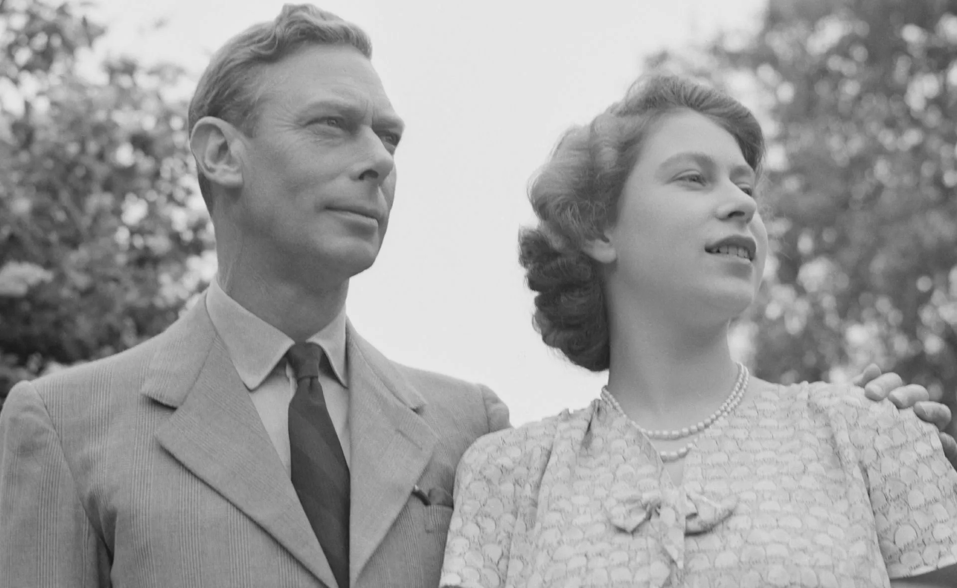 Father and daughter stand together, looking into the distance, against a backdrop of trees.