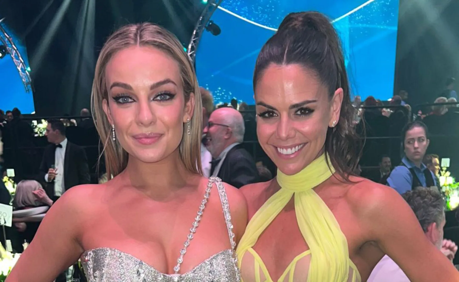 Two women in elegant dresses smiling at a formal event, with gala attendees and a stage in the background.