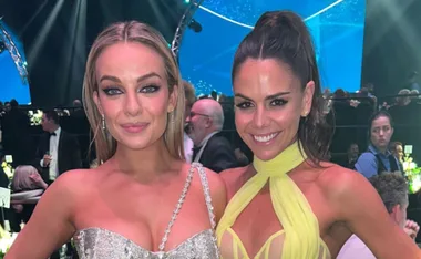 Two women in elegant dresses smiling at a formal event, with gala attendees and a stage in the background.
