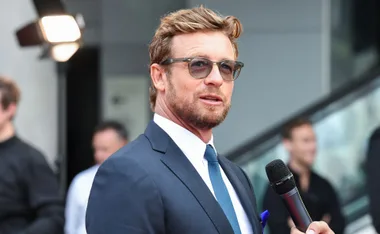 Man in sunglasses holding a microphone, dressed in a suit, speaking at an event outdoors.