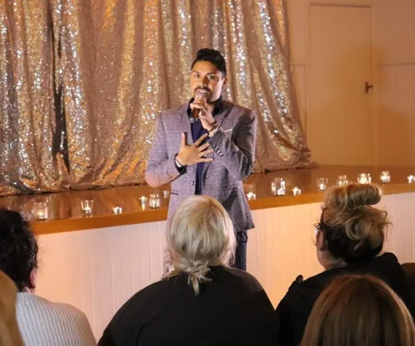 Male speaker in a blazer performs on stage with candles, addressing a seated audience in a dimly lit room.